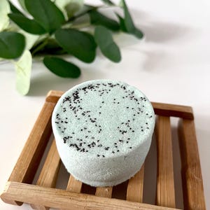 May include: A round, pale mint green bath bomb with a textured surface speckled with small black particles, resting on a slatted wooden soap dish. The bath bomb has a cylindrical shape with a flat top and sides, showcasing a matte finish.