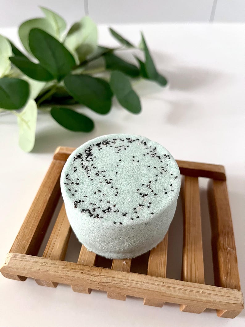 May include: A round, pale mint green bath bomb with a textured surface speckled with small black particles, resting on a slatted wooden soap dish. The bath bomb has a cylindrical shape with a flat top and sides, showcasing a matte finish.