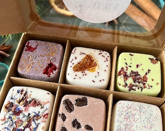 May include: A cardboard box containing six square bath bombs. The top left is purple with red petals, top middle is white with an orange slice, top right is light green with dried flowers, bottom left is white with colorful petals, bottom middle is tan with coffee beans, and bottom right is white with glitter.