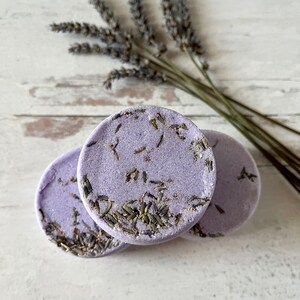 May include: Three round lavender bath bombs with dried lavender flowers embedded in the surface. The bath bombs are on a white wooden surface.