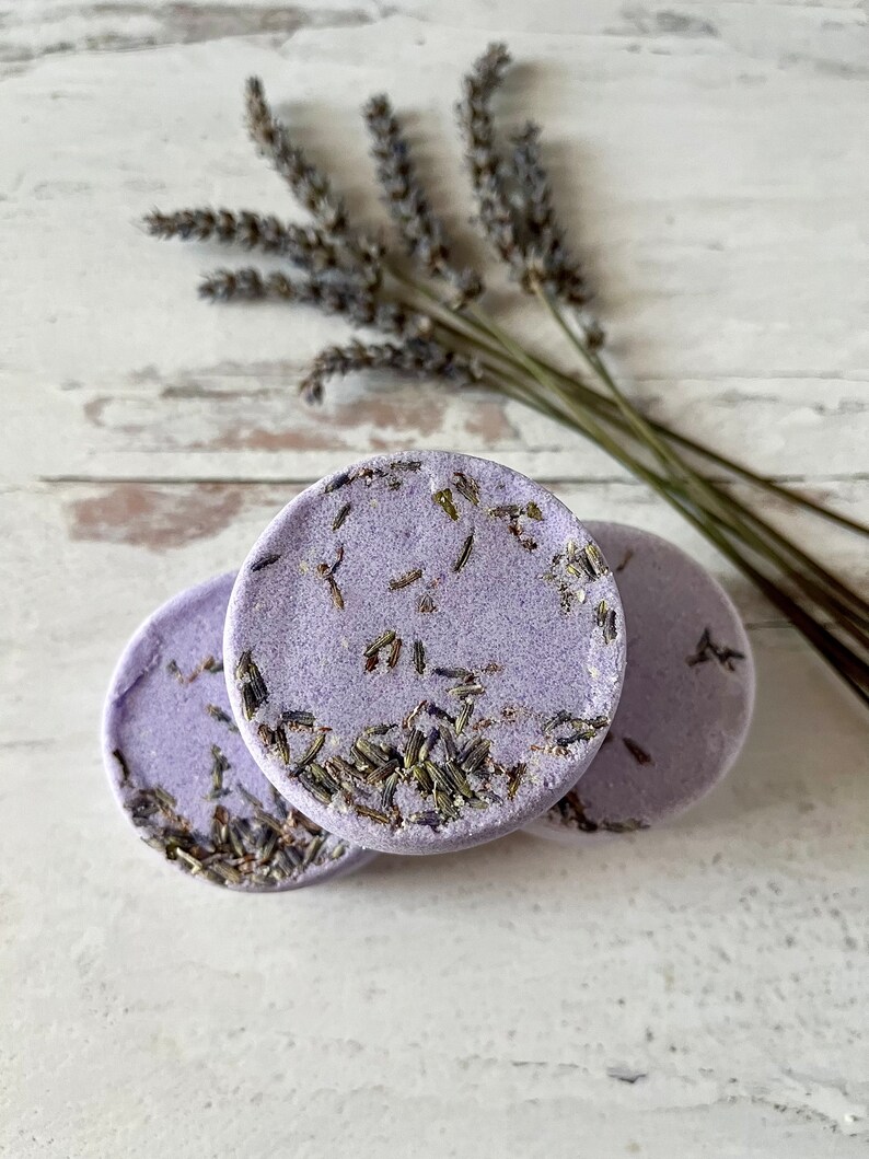 May include: Three round lavender bath bombs with dried lavender flowers embedded in the surface. The bath bombs are on a white wooden surface.