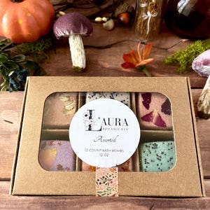 May include: A rectangular brown cardboard box with a clear window containing twelve square bath bombs in various colors and textures. A white circular label on the front reads Laura Botanicals, Assorted, 12 Count Bath Bombs, 12 oz.