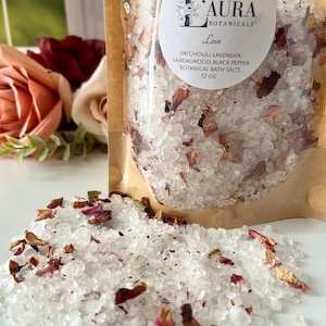 May include: A 12 oz pouch of L'Aura Botanicals Love botanical bath salts containing white salt crystals mixed with dried flower petals. The label lists patchouli, lavender, sandalwood, and black pepper as ingredients.