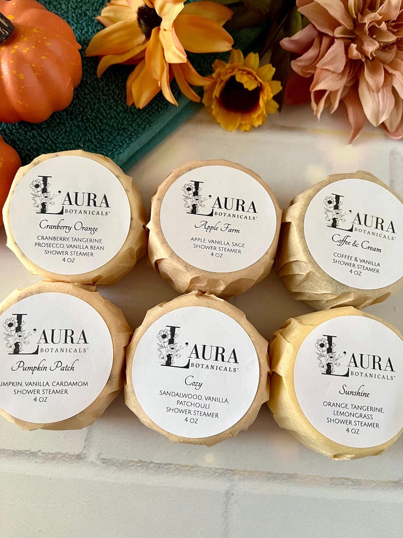 May include: Six round shower steamers wrapped in tan paper, each with a white label reading Laura Botanicals. Scents include Cranberry Orange, Apple Farm, Coffee & Cream, Pumpkin Patch, Cozy, and Sunshine. Each label specifies a 4 oz weight.