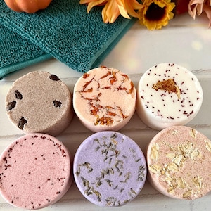 May include: Six round bath bombs arranged in two rows. The top row features a brown bomb with dark inclusions, a peach bomb with orange petals, and a white bomb with red specks. The bottom row includes a pink bomb, a purple lavender-topped bomb, and a tan oat-topped bomb.