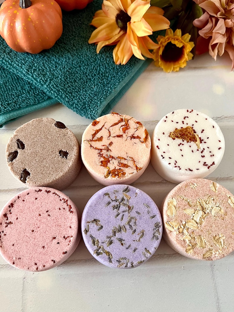 May include: Six round bath bombs arranged in two rows. The top row features a brown bomb with dark inclusions, a peach bomb with orange petals, and a white bomb with red specks. The bottom row includes a pink bomb, a purple lavender-topped bomb, and a tan oat-topped bomb.