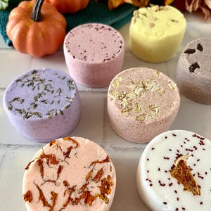 May include: A collection of seven round, cylindrical bath bombs in various colors including lavender, pink, peach, white, and yellow. Each bath bomb features unique botanical inclusions such as dried flower petals, lavender buds, and oats.