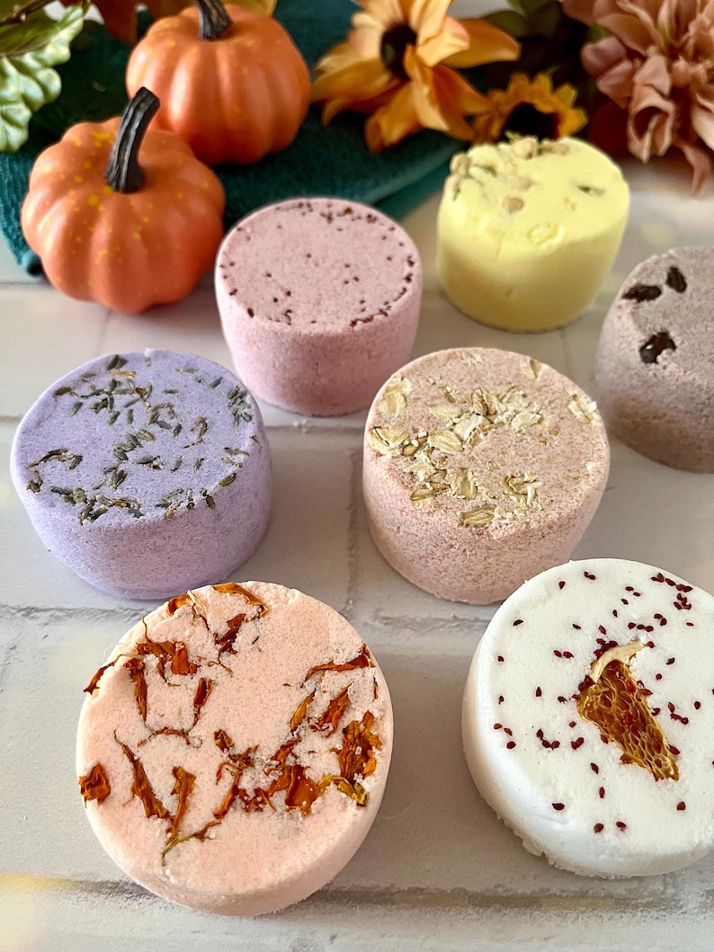 May include: A collection of seven round, cylindrical bath bombs in various colors including lavender, pink, peach, white, and yellow. Each bath bomb features unique botanical inclusions such as dried flower petals, lavender buds, and oats.