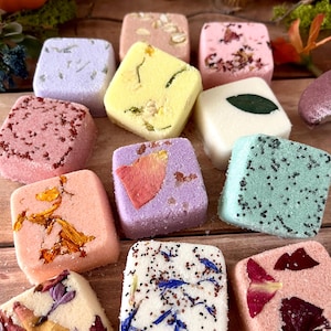 May include: A collection of square-shaped bath bombs in various colors including yellow, pink, purple, green, and white. Each bath bomb is decorated with dried flower petals, leaves, or small seeds embedded on the top surface of the compressed powder.