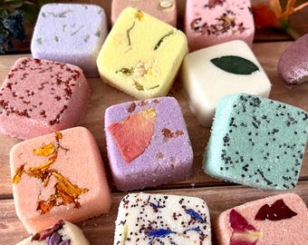 May include: A collection of square-shaped bath bombs in various colors including yellow, pink, purple, green, and white. Each bath bomb is decorated with dried flower petals, leaves, or small seeds embedded on the top surface of the compressed powder.
