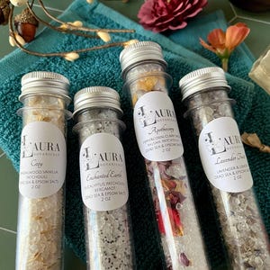May include: Four clear glass test tubes filled with bath salts, each featuring a white label with the brand name Laura Botanicals. The labels list the scents Cozy, Enchanted Earth, Peppercorn Clary Sage, and Lavender Tea, each containing 2 oz of salts.