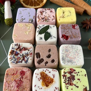 May include: A collection of twelve square bath bombs in various colors, including lavender, pink, yellow, white, and green. Each bath bomb is decorated with dried flower petals, herbs, coffee beans, or citrus slices, arranged in a grid on a green surface.