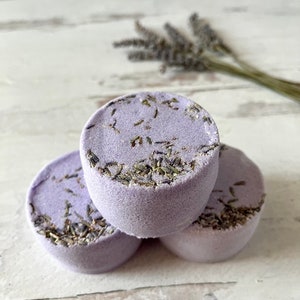 May include: Three lavender bath bombs with dried lavender buds on top. The bath bombs are stacked on top of each other on a white wooden surface.