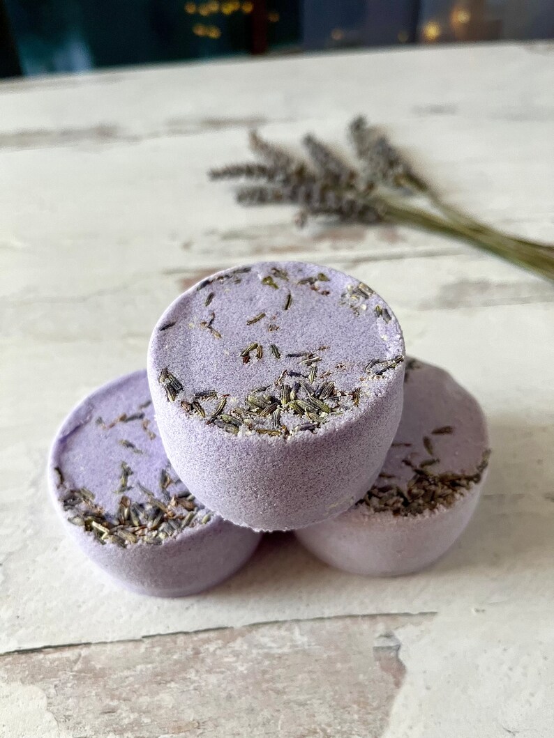 May include: Three lavender bath bombs with dried lavender buds on top. The bath bombs are stacked on top of each other on a white wooden surface.