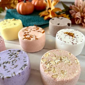 May include: A collection of seven cylindrical bath bombs in various colors and textures. The set includes a purple lavender-topped bomb, a pink oat-topped bomb, a yellow bomb, a peach floral-topped bomb, a white bomb with red specks, and a brown bomb.