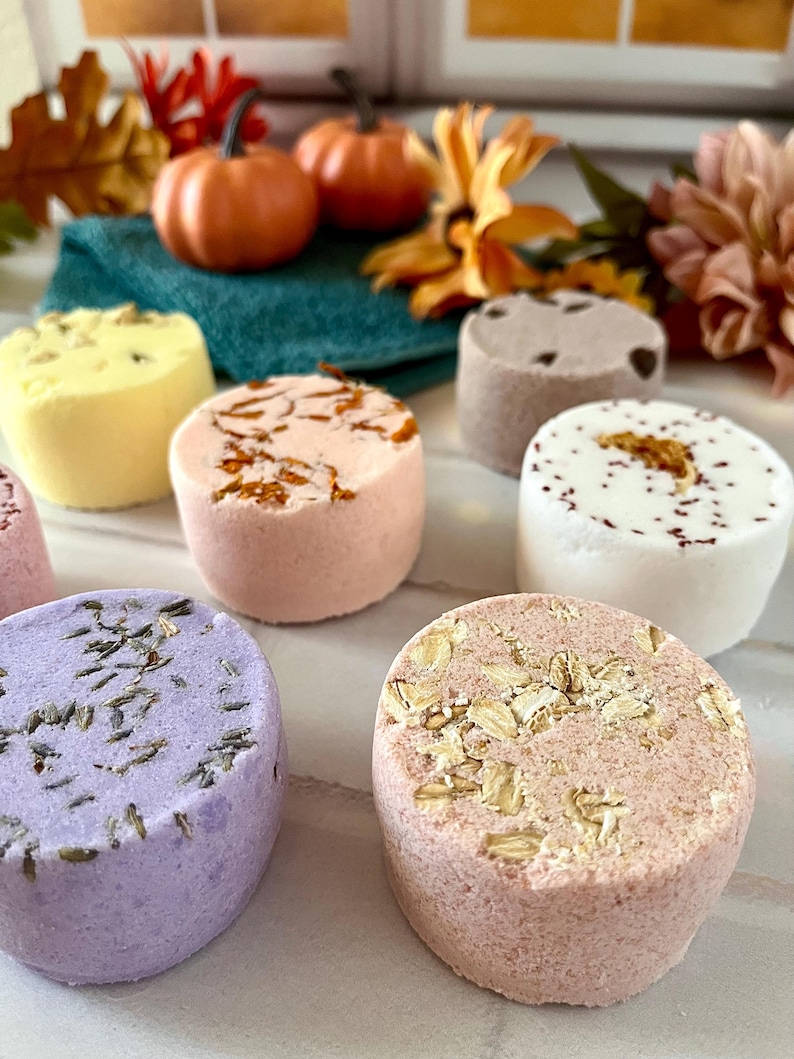 May include: A collection of seven cylindrical bath bombs in various colors and textures. The set includes a purple lavender-topped bomb, a pink oat-topped bomb, a yellow bomb, a peach floral-topped bomb, a white bomb with red specks, and a brown bomb.