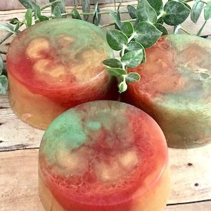 May include: Three round bars of handmade soap with a swirl of green, orange, and red colors. The soap is sitting on a wooden surface with green leaves.