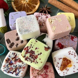 May include: A collection of square-shaped bath bombs in various colors, including white, pink, green, and purple. The bath bombs are decorated with dried flower petals, orange slices, coffee beans, oats, and glitter, arranged with cinnamon sticks and star anise.