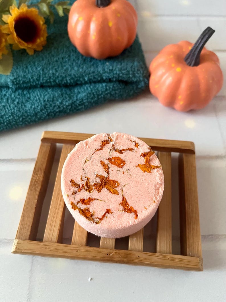 May include: A round, light pink bath bomb embedded with dried orange flower petals, resting on a rectangular wooden slatted soap dish. The bath bomb has a textured surface and is positioned in front of a dark teal towel and two small orange pumpkins.