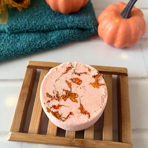 May include: A round, light pink bath bomb embedded with dried orange flower petals, resting on a rectangular wooden slatted soap dish. The bath bomb has a textured surface and is positioned in front of a dark teal towel and two small orange pumpkins.