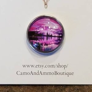 May include: A round, silver-colored, glass-like button with a pink and purple mountain scene reflected in a lake. The button is on a white card with the text "Camo and Ammo Boutique" and a website address.