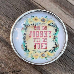 May include: A round, silver-framed charm with a white background and a floral design. The charm features a vintage-style sign with the text "You Be Johnny I'll Be June".