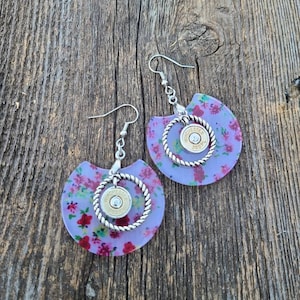 May include: A pair of earrings with a floral design and a silver ring with a bullet casing in the center. The earrings are on a wooden background.