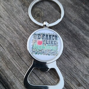 May include: Silver bottle opener keychain with a round, white, and green Applebee's logo that says "We fancy like Applebee's on a date night".