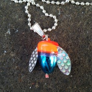 May include: A silver chain necklace with a blue, orange, and iridescent lure pendant. The lure has two iridescent wings and a red bead at the bottom.