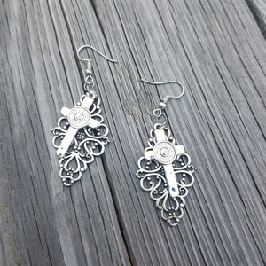 May include: Silver filigree earrings with a bullet casing cross charm. The earrings are dangling and have a hook closure.