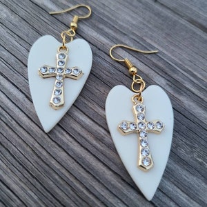 May include: A pair of white heart-shaped earrings with gold cross charms adorned with clear rhinestones.