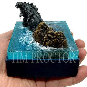May include: A miniature Godzilla figure standing on a rocky island in a blue resin base. The base has a clear resin top and a black wooden bottom. The text "TIM PROCTOR" is printed on the base.