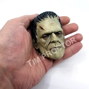 May include: A detailed, hand-painted Frankenstein monster head figurine. The figure has a pale green complexion, black hair, and visible stitches and scars. The text "TIM PROCTOR" is visible on the image.