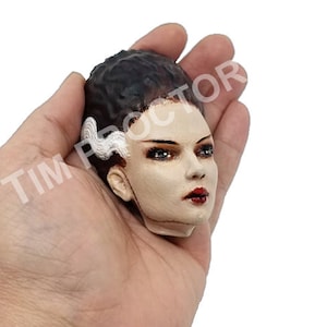 May include: A sculpted head of a female figure with black hair, white makeup, and red lips. The figure has a scar across her forehead and a white scar on the side of her head. The head is held in a hand.