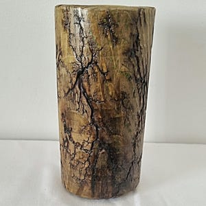 Op de afbeelding: Een cilindrische houten vaas met een rustieke bruine en groene afwerking. De vaas heeft een uniek gebarsten en getextureerd oppervlak, waardoor hij er natuurlijk en organisch uitziet.