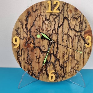 May include: A wooden clock with a dark brown wood grain and a black and green clock face. The clock face has yellow numbers and a black hour hand and a green minute hand. The clock is on a clear plastic stand.