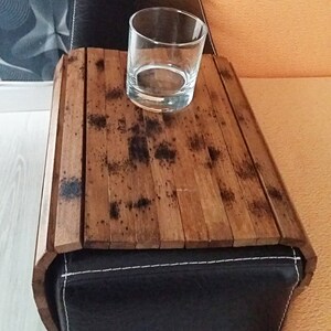 May include: A wooden tray with a dark brown finish, designed to fit over the arm of a couch or chair. The tray has a glass on top, suggesting it can be used to hold drinks or snacks.