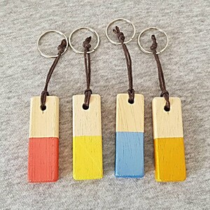 Puede incluir: Cuatro llaveros de madera con diferentes pinturas de colores en la mitad inferior. Los llaveros son rojo, amarillo, azul y dorado.