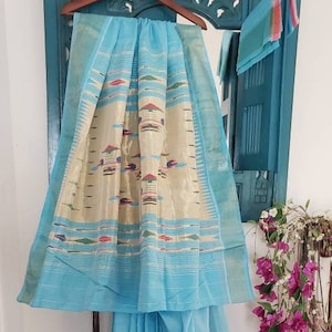 May include: A light blue saree with a cream and gold patterned border. The saree has a light blue pallu with a cream and gold patterned border. The saree is draped over a hanger and is displayed against a blue background.