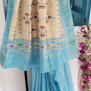 May include: A light blue saree with a cream and blue patterned border. The saree has a cream colored pallu with a geometric design in blue, red, and green.