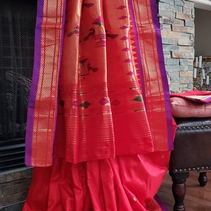 May include: A red and gold saree with a purple border. The saree is draped over a wooden stool and has a patterned design.
