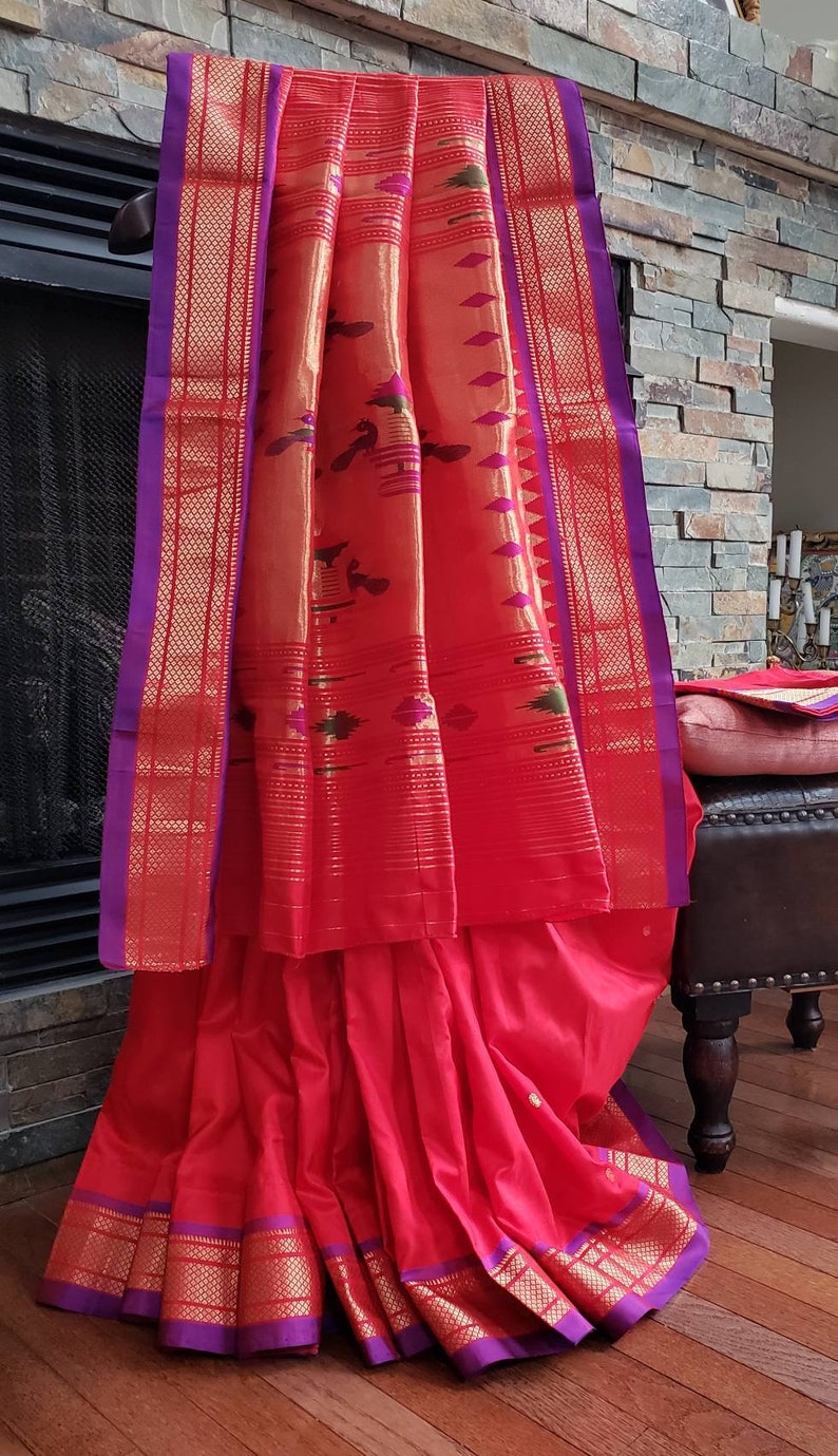 May include: A red and gold saree with a purple border. The saree is draped over a wooden stool and has a patterned design.