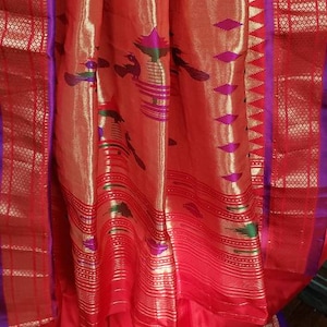 May include: A red and gold silk saree with a purple border and intricate patterns. The saree is draped over a wooden surface.