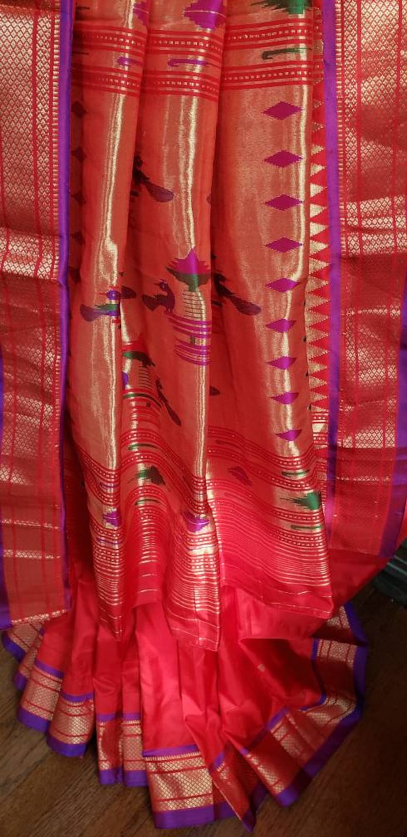 May include: A red and gold silk saree with a purple border and intricate patterns. The saree is draped over a wooden surface.