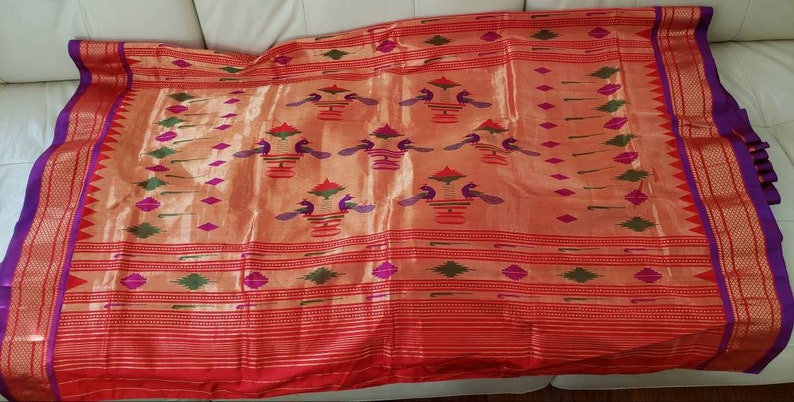 May include: A red and gold silk saree with a purple border and intricate peacock designs. The saree has a traditional Indian design with a woven pattern.