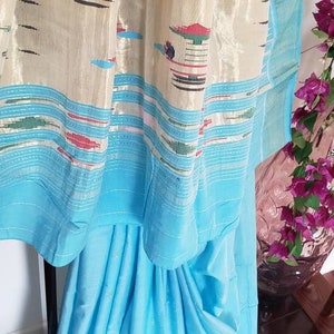 May include: A light blue saree with a cream colored border featuring a geometric pattern. The border has a silver shimmer and a repeating pattern of small, colorful figures.