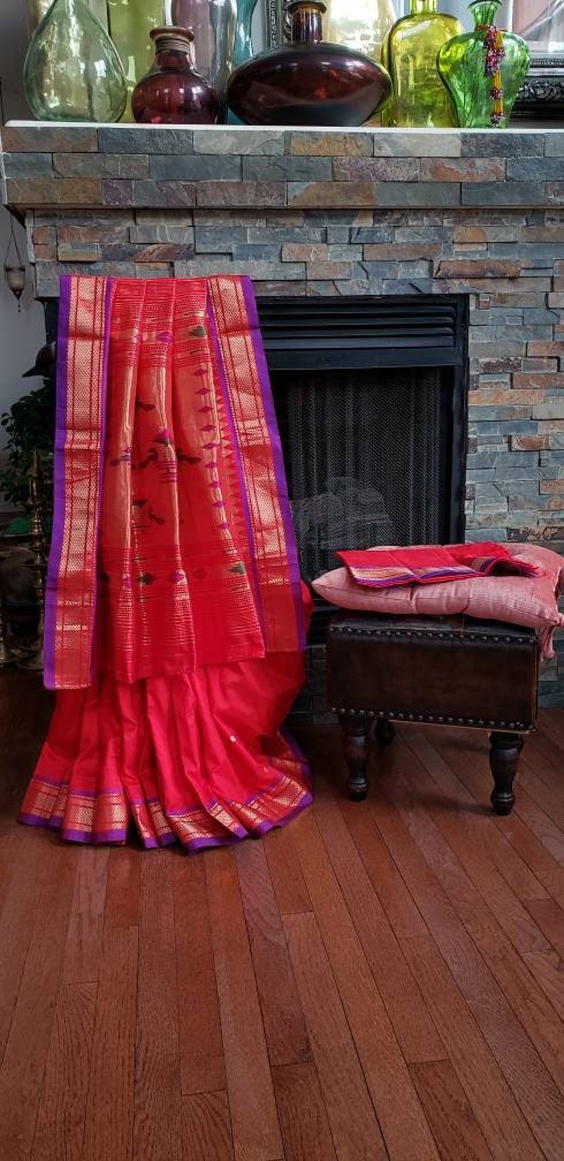 May include: A red and gold saree with purple trim, draped over a wooden stool with a pink cushion. The saree is decorated with intricate patterns and designs.