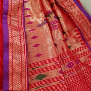 May include: A red and gold saree with a purple border and intricate woven patterns. The saree is made of silk and has a soft, luxurious feel.