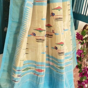 May include: A light blue saree with a cream and gold patterned border. The saree has a light blue pallu and a cream and gold body with colorful geometric designs.