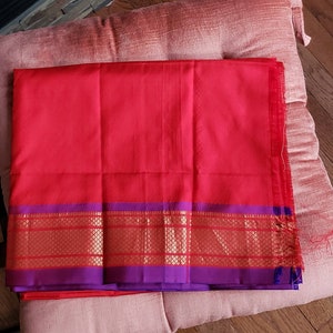 May include: A red and purple silk saree with a gold border. The saree is folded and lying on a pink cushion.